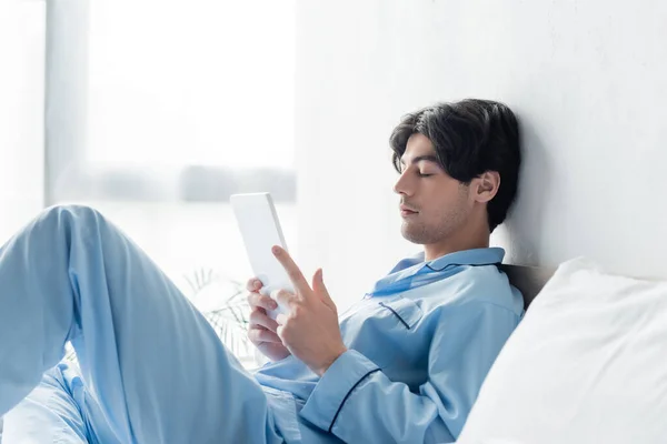 Joven con los ojos cerrados sentado en la cama con la tableta digital - foto de stock