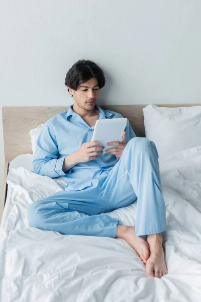 Junger Mann barfuß im blauen Pyjama mit digitalem Tablet im Schlafzimmer — Stockfoto