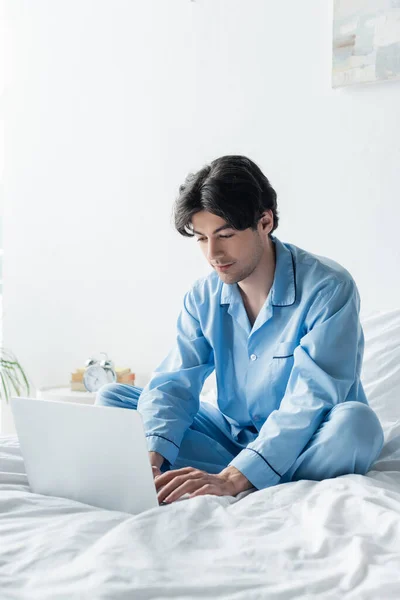 Mann im blauen Schlafanzug sitzt mit überkreuzten Beinen im Bett und tippt auf Laptop — Stockfoto