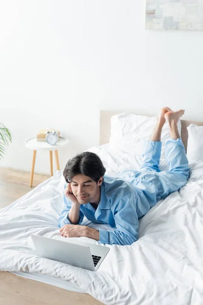 Uomo sorridente su pigiama blu guardare film sul computer portatile sul letto — Foto stock