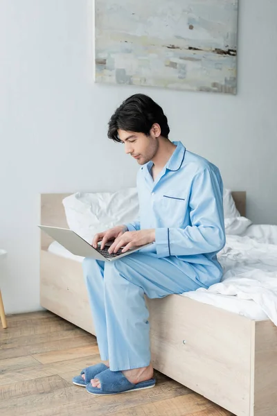 Mann in blauem Schlafanzug und Hausschuhen sitzt auf Bett und tippt auf Laptop — Stockfoto