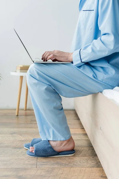 Vista recortada del hombre en pijama azul y zapatillas de escribir en el ordenador portátil en el dormitorio - foto de stock