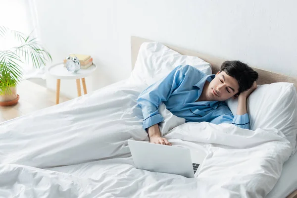 Hombre positivo en pijama azul viendo la película en el ordenador portátil en la cama - foto de stock