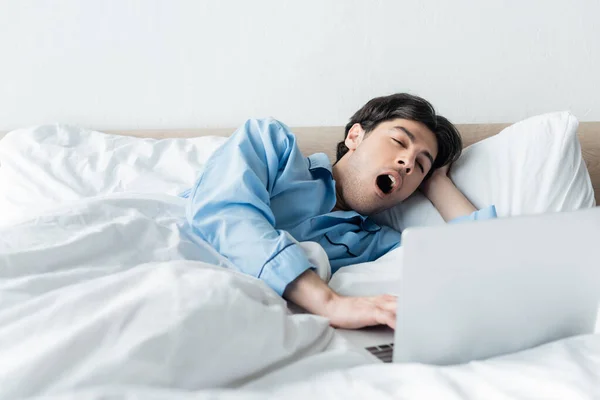 Hombre soñoliento bostezando cerca de la computadora portátil mientras está acostado en la cama por la mañana - foto de stock