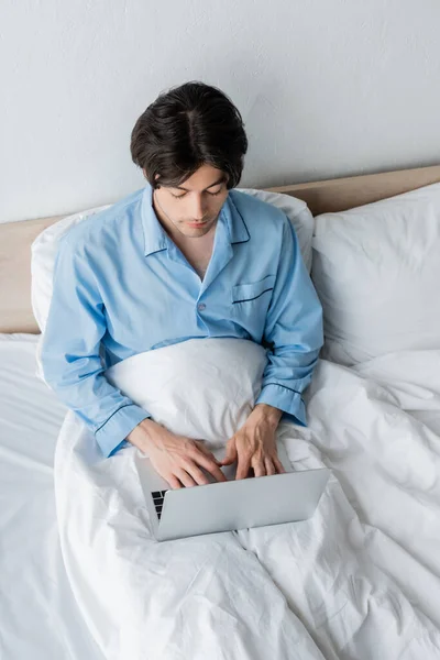 Visão de alto ângulo do homem em pijama azul digitando no laptop enquanto sentado na cama — Fotografia de Stock