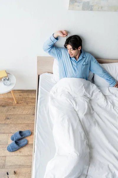 Hochwinkelaufnahme des Mannes im blauen Schlafanzug, der sich im Bett in der Nähe des Vintage-Weckers auf dem Nachttisch ausbreitet — Stockfoto