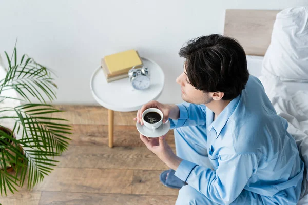 Vue grand angle de l'homme en pyjama bleu assis sur le lit avec une tasse de café — Photo de stock