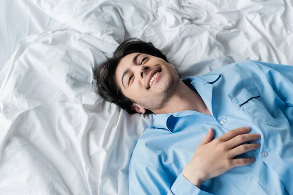 Top view of happy brunette man in blue pajamas lying on white bedding — Stock Photo