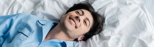 Vue de dessus du jeune homme brune souriant tout en étant couché sur la literie blanche, bannière — Photo de stock