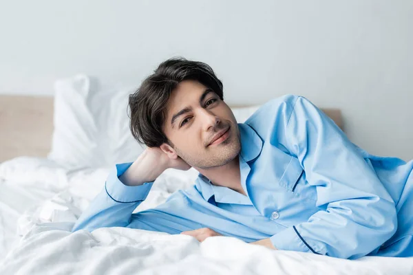 Pleased man in blue pajamas looking at camera while lying on white bedding — Stock Photo