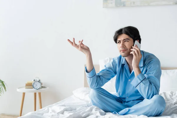 Вдумливий чоловік сидить на ліжку з схрещеними ногами і жестикулює під час розмови на смартфоні — Stock Photo