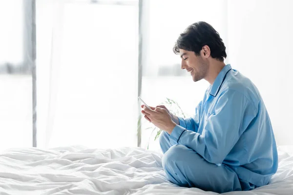 Seitenansicht eines lächelnden Mannes, der mit überkreuzten Beinen auf dem Bett sitzt und mit dem Handy chattet — Stockfoto