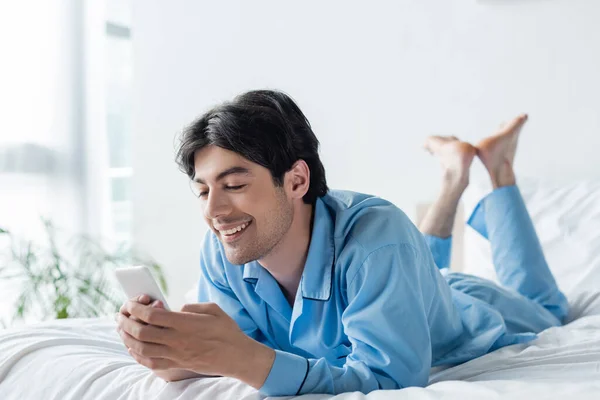 Cheerful barefoot man in blue pajamas messaging on mobile phone in bedroom — Stock Photo