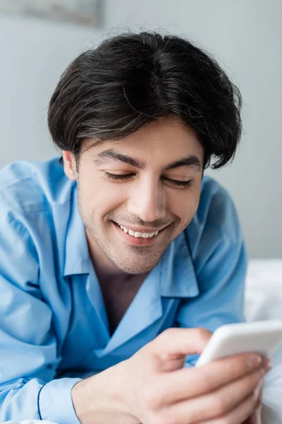 Vista de cerca del hombre alegre en pijama azul usando el teléfono móvil en el dormitorio - foto de stock