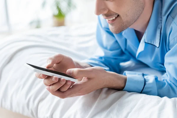 Vista ritagliata di uomo sorridente messaggistica sul telefono cellulare sul letto — Foto stock