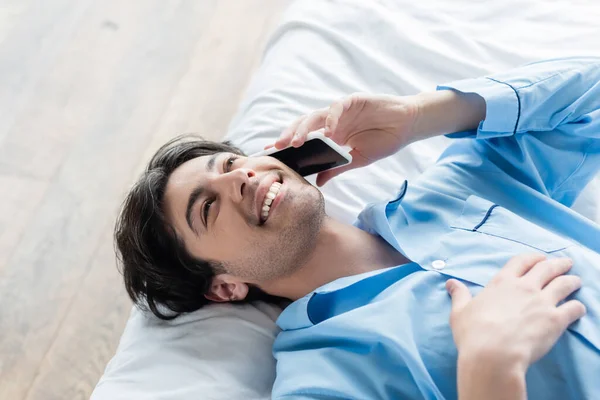 Blick von oben auf einen glücklichen Mann im blauen Pyjama, der auf dem Bett liegt und mit dem Handy telefoniert — Stockfoto