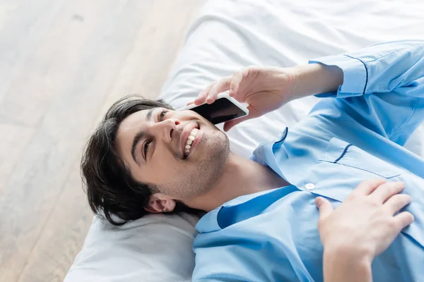 Vista aérea del hombre feliz en pijama azul acostado en la cama y hablando por teléfono móvil - foto de stock