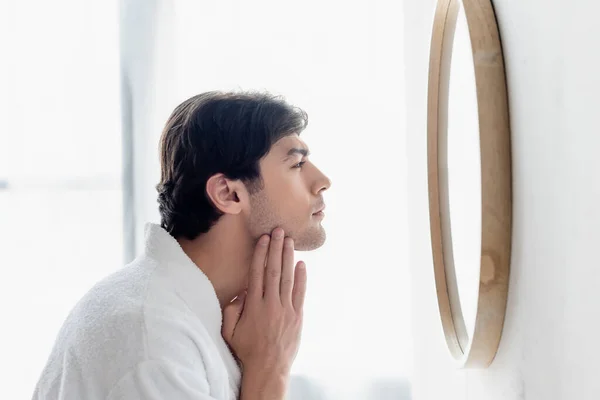 Side view of young man in bathrobe touching face in bathroom — Stock Photo