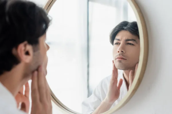 Брюнетка людина торкається обличчя, дивлячись у дзеркало ванної кімнати — стокове фото