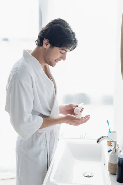 Vista lateral do homem feliz em roupão de banho aplicando creme de mão perto de produtos de higiene pessoal na pia — Fotografia de Stock