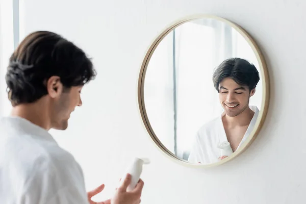 Uomo sorridente in accappatoio bianco che tiene la crema per le mani vicino allo specchio in bagno — Foto stock