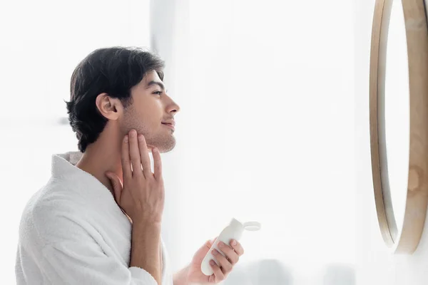 Vue latérale de l'homme brunette appliquant du lait cosmétique sur le visage dans la salle de bain — Photo de stock