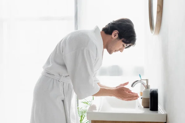 Vista lateral del joven en albornoz blanco cerca del lavabo y artículos de tocador en el baño - foto de stock