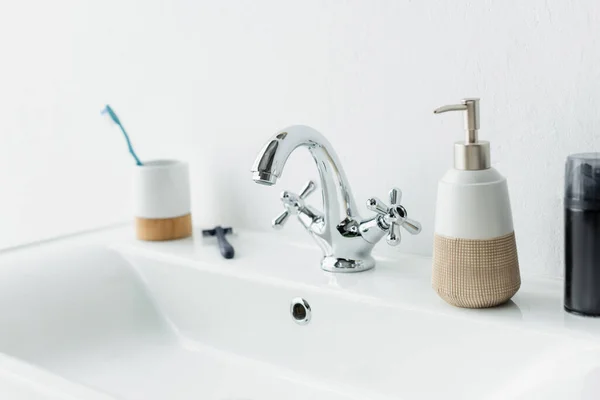 Shaving foam, liquid soap, safety razor and toothbrush on white sink with faucet — Stock Photo