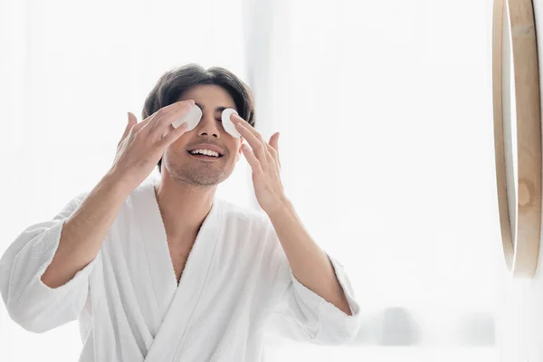 Hombre alegre en albornoz blanco limpiando los ojos con almohadillas de algodón en el baño - foto de stock