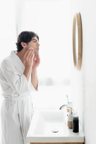 Side view of man in white bathrobe cleaning face with cotton pad in bathroom — Stock Photo