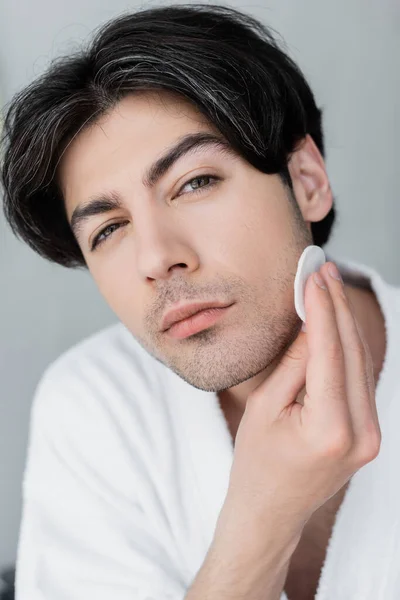 Young man looking at camera while wiping face with cotton pad isolated on grey — Stock Photo