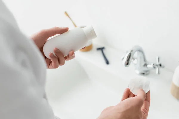 Vista recortada del hombre sosteniendo contenedor cosmético y almohadilla de algodón cerca del fregadero borroso - foto de stock