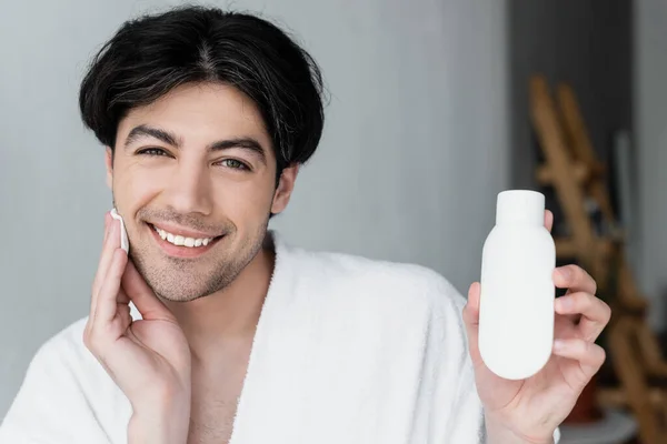 Felice uomo che tiene il contenitore con latte cosmetico mentre pulisce il viso con batuffolo di cotone — Foto stock
