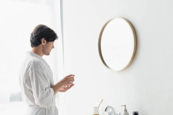 Hombre joven en albornoz blanco celebración de crema cosmética cerca del espejo en el baño - foto de stock