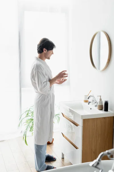 Vue pleine longueur de l'homme en peignoir blanc tenant crème cosmétique près du miroir et articles de toilette sur l'évier — Photo de stock