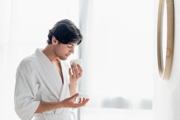 Hombre joven en albornoz oliendo crema cosmética en el baño - foto de stock