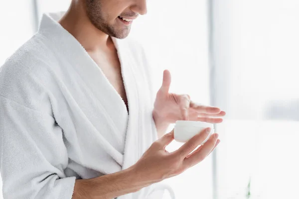 Vista cortada do homem em branco roupão segurando recipiente com creme cosmético — Fotografia de Stock