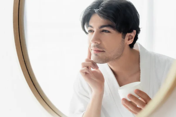 Brünetter Mann im Bademantel schaut in Spiegel und trägt Gesichtscreme auf — Stockfoto