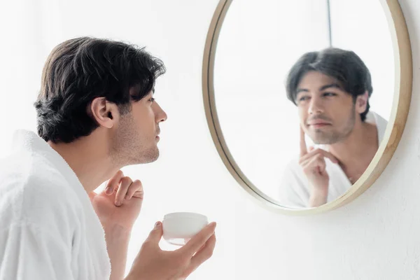 Morena positiva hombre aplicando crema facial cerca del espejo en el baño - foto de stock