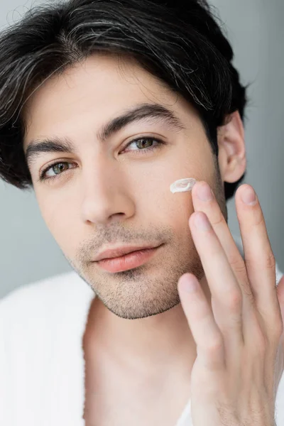 Vue rapprochée du jeune homme qui applique de la crème pour le visage et regarde la caméra — Photo de stock