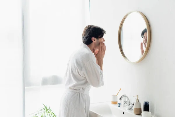 Brunette homme en peignoir blanc appliquant crème visage près du miroir et de l'évier — Photo de stock