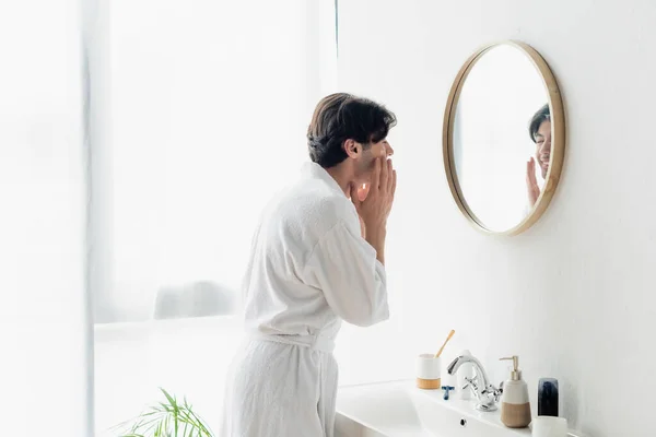 Homem morena em roupão branco aplicando creme facial perto do espelho e pia — Fotografia de Stock