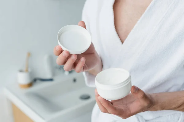 Vista cortada do homem em roupão de banho branco segurando creme cosmético no banheiro desfocado — Fotografia de Stock