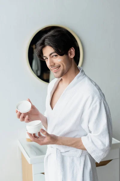 Man in bathrobe smiling at camera while holding cosmetic cream in bathroom — Stock Photo