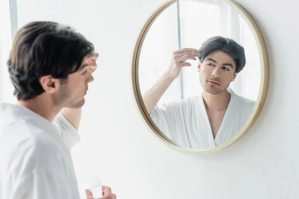 Jeune homme en peignoir blanc appliquant sérum cosmétique près du miroir dans la salle de bain — Photo de stock