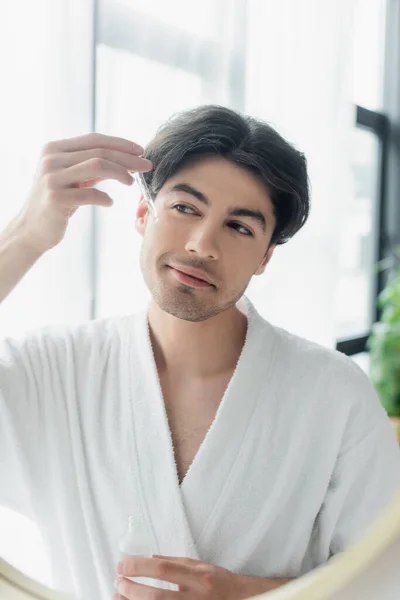 Homem morena em roupão de banho branco sorrindo ao aplicar soro cosmético perto do espelho — Fotografia de Stock