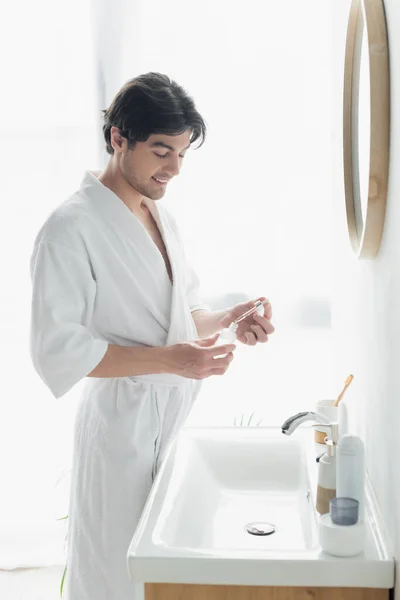 Hombre sonriente de albornoz blanco sosteniendo la pipeta y la botella con suero cosmético - foto de stock