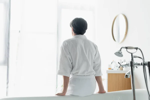 Back view of brunette man in white bathrobe sitting on bathtub — Stock Photo