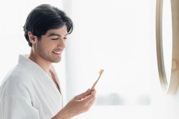 Glücklicher Mann im weißen Bademantel mit Blick auf die Zahnbürste im Badezimmer — Stockfoto