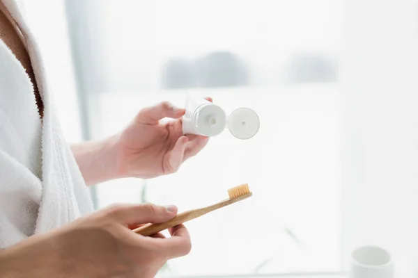 Vista recortada del hombre en albornoz blanco sosteniendo cepillo de dientes y tubo de pasta de dientes - foto de stock